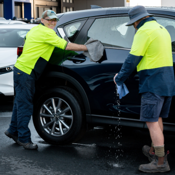 Two workers in hi-vis shirts washing a dark SUV in a car park.